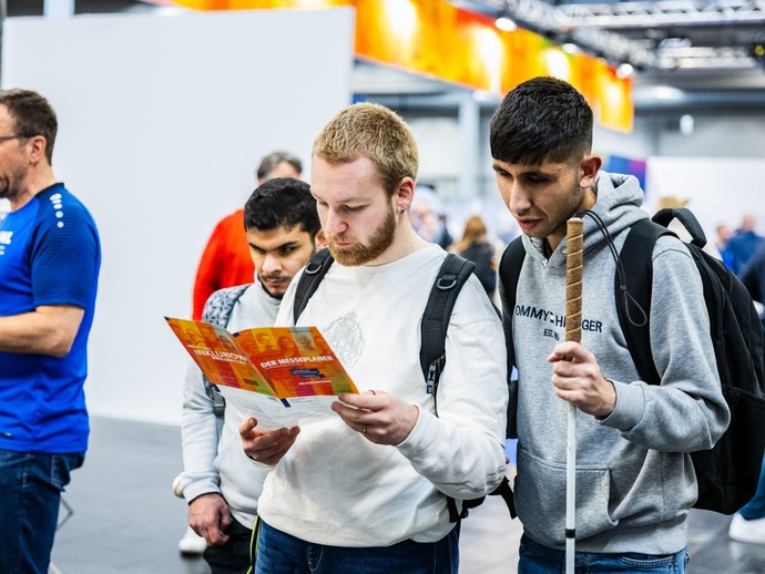 Zwei junge Männer betrachten gemeinsam einen Messeplaner. (vergrößerte Bildansicht wird geöffnet)