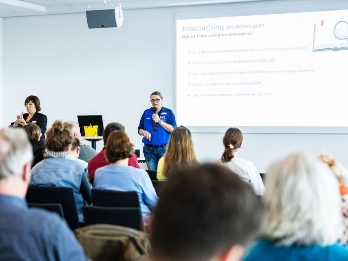 Eine Frau hält einen Vortrag mit Mikrofon, während Zuhörende im Bild zu sehen sind und eine Gebärdendolmetscherin übersetzt. (vergrößerte Bildansicht wird geöffnet)