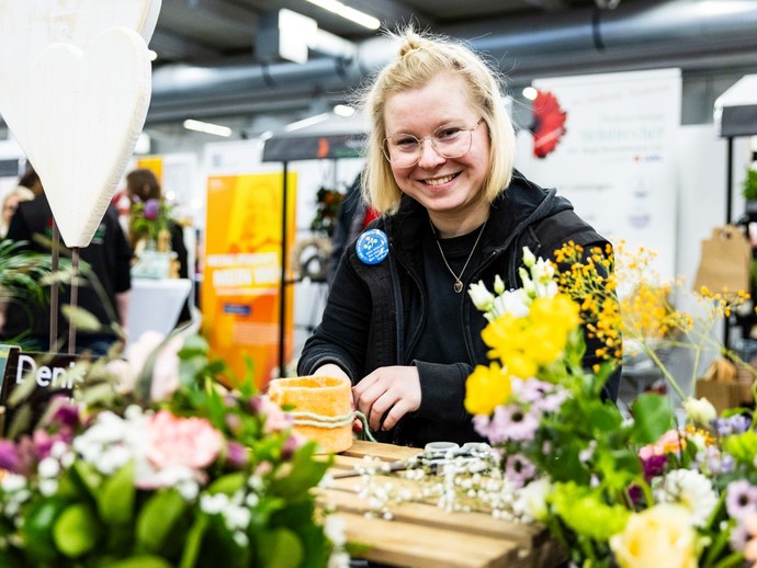 Eine Frau lächelt in die Kamera, während sie von Blumen umgeben ist. (vergrößerte Bildansicht wird geöffnet)