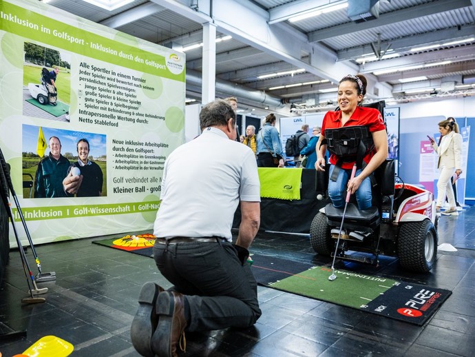 Eine Frau im Rollstuhl hält einen Minigolfschläger, während ein Mann ihr gegenüber etwas erklärt. (vergrößerte Bildansicht wird geöffnet)