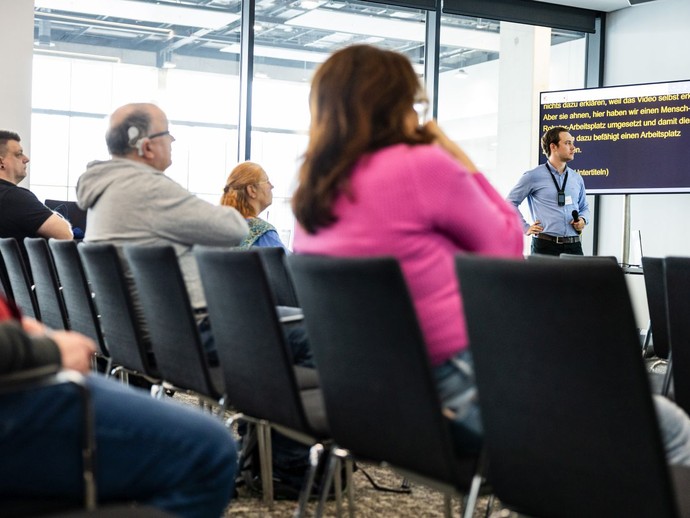 Ein Mann hält in einem Seminarraum einen Vortrag mit Mikrofon, während Zuhörende im Vordergrund zu sehen sind und ein Bildschirm mit Schriftdolmetschu (vergrößerte Bildansicht wird geöffnet)