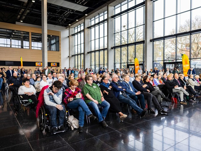 Viele Besucherinnen und Besucher sitzen auf Stuhlreihen oder stehen dahinter und hören aufmerksam zu. (vergrößerte Bildansicht wird geöffnet)