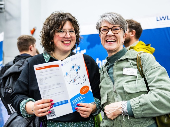 Zwei Frauen lächeln in die Kamera, eine von ihnen hält eine Broschüre in der Hand. (vergrößerte Bildansicht wird geöffnet)