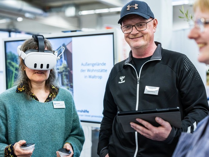 Eine Frau trägt eine VR-Brille und hält zwei Controller, während ein Mann daneben ein Tablet in der Hand hält. (vergrößerte Bildansicht wird geöffnet)