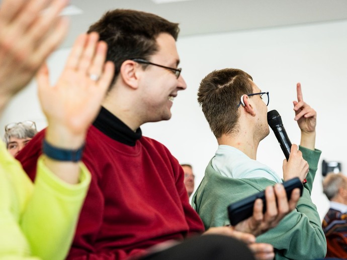 Ein Mann meldet sich in einem Seminarraum, hält ein Mikrofon und stellt eine Frage. (vergrößerte Bildansicht wird geöffnet)