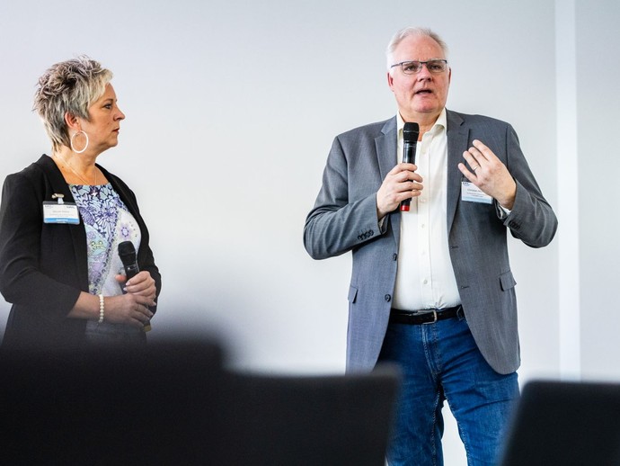 Ein Mann spricht in ein Mikrofon, während eine Frau daneben steht, ebenfalls ein Mikrofon hält und zuhört. (vergrößerte Bildansicht wird geöffnet)