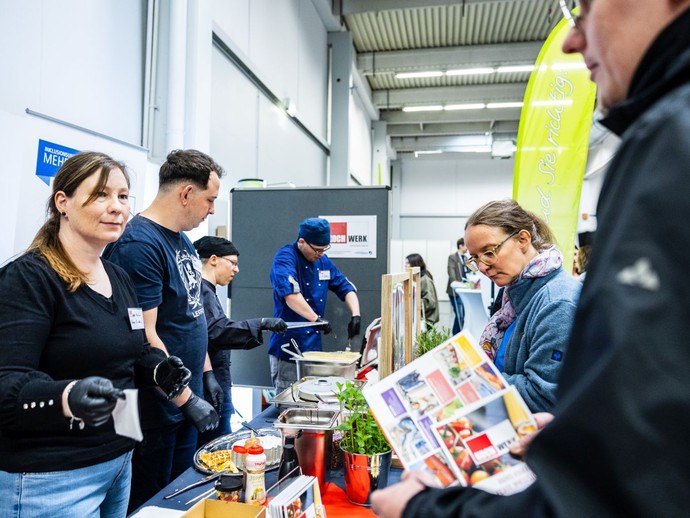 Zwei Personen kaufen Waffeln, während auf der anderen Seite mehrere Personen Speisen ausgeben. (vergrößerte Bildansicht wird geöffnet)