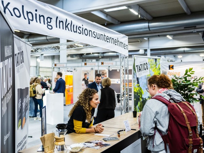 Eine Frau informiert sich an einem Messestand, während eine andere Person ihr etwas erklärt. (vergrößerte Bildansicht wird geöffnet)